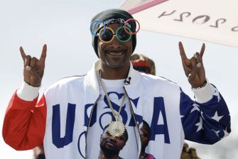 Snoop Dogg wearing Olympic rings glasses and a USA-themed jacket, posing with a playful gesture.