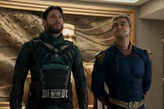 Two characters from a superhero series stand in a stylized interior, one wearing a green suit with armor and the other in a blue and gold costume, both with confident expressions.