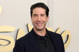 David Schwimmer smiling at an event, wearing a dark blazer over a black shirt, with a branded background.