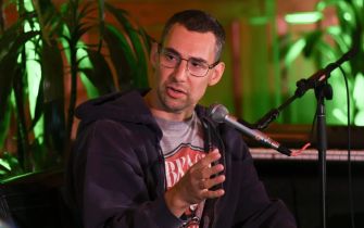 A man speaking into a microphone while seated, surrounded by greenery, during a discussion or interview.