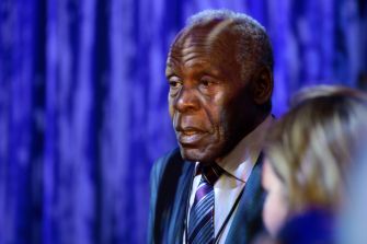 A man in formal attire appears contemplative, standing against a backdrop of blue curtains.