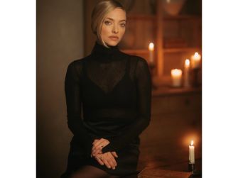 Amanda Seyfried posing in a dimly lit setting with candles, wearing a black turtleneck and looking contemplative.
