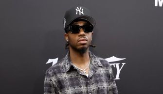 Metro Boomin at a public event, wearing sunglasses and a baseball cap, with a checkered shirt in a stylish pose.