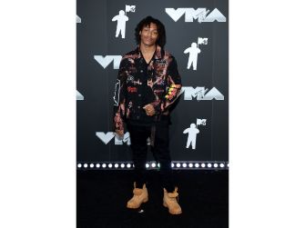 A young man posing at an event with a black and colorful jacket, black pants, and tan boots.