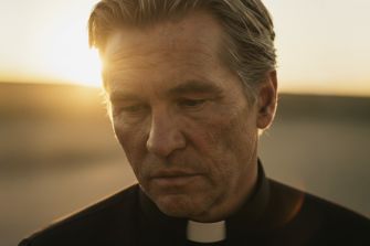 A solemn-looking man dressed in a priest's collar, gazing downward against a sunset backdrop.