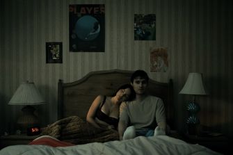 A scene from the horror film "Obsession," featuring two characters sitting on a bed in a dimly lit bedroom.