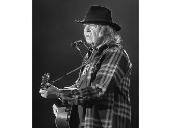Neil Young performing on stage with a guitar, wearing a plaid shirt and hat.