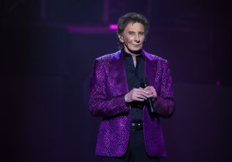 Barry Manilow performing on stage in a purple suit, holding a microphone.