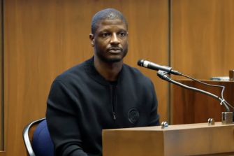 A man testifying in a courtroom setting, seated at a witness stand with a microphone in front of him.