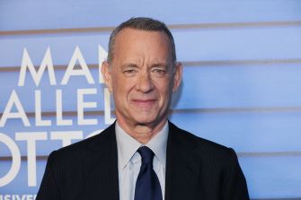 Tom Hanks at a film event, wearing a dark suit and tie, smiling at the camera.