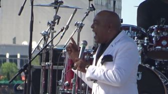 A singer performing on stage in a white suit, with drums visible in the background.