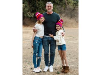 A man stands in an outdoor setting with two young girls, all three wearing colorful beanie hats and smiling for the camera.