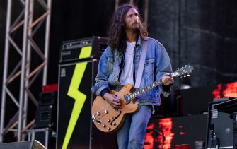 A guitarist performing on stage with a guitar, dressed in a denim jacket, with band equipment and lights in the background.