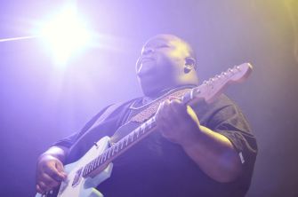 A musician performing on stage with a guitar, illuminated by bright stage lights.
