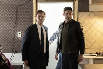 Two male characters positioned in an interior setting, one in a suit and the other in a leather jacket, appearing alert and engaged in a scene from a drama series.