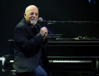 Billy Joel smiling while performing at a piano during a live concert.