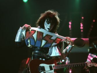 A musician on stage holding a banner with the band name "KISS" while playing an electric guitar.