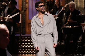 Bad Bunny smiling and performing during his debut on Saturday Night Live, wearing sunglasses and a light gray outfit.