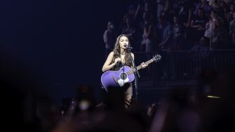 Olivia Rodrigo performing on stage with a purple guitar and a cheering audience in the background.