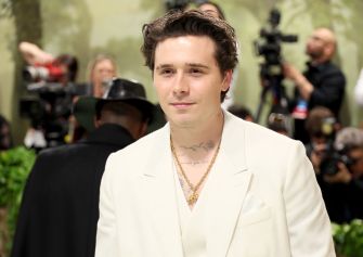 Brooklyn Beckham at a public event, wearing a white suit and gold accessories, with photographers in the background.