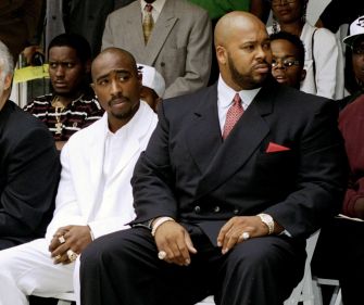 Tupac Shakur seated next to Suge Knight at a public event, with a crowd in the background.