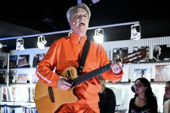 David Byrne performing on stage with a guitar, wearing an orange outfit, during a concert.