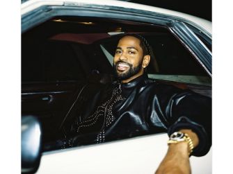 Big Sean smiling while seated in a car, wearing a black leather jacket and a watch.