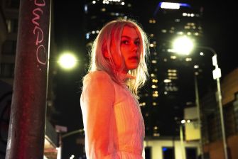 A woman with long, light-colored hair stands under streetlights in an urban environment at night, with city buildings illuminated in the background.