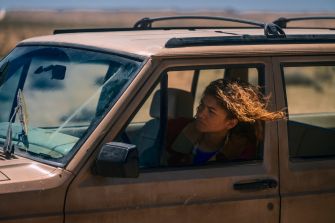 A character sitting in a vehicle, looking contemplative, with wind-blown hair.